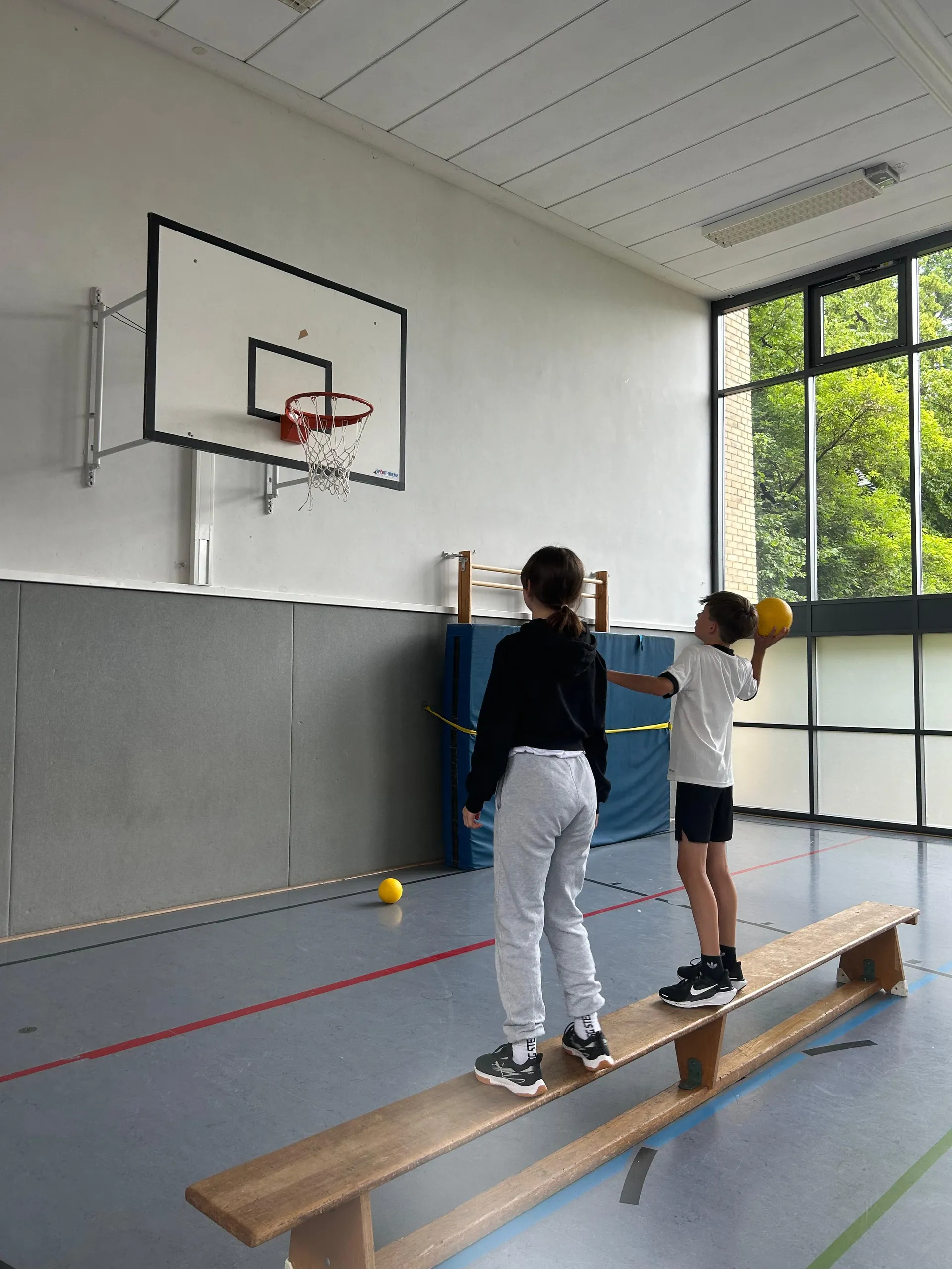 Zwei Kinder stehen auf einer Holzbank in einer Sporthalle, eines hält einen gelben Ball.