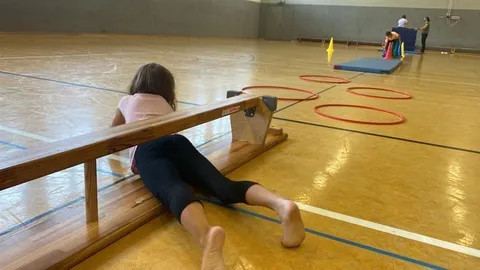 Ein Kind liegt in einer Sporthalle neben einer niedrigen Holzbank.