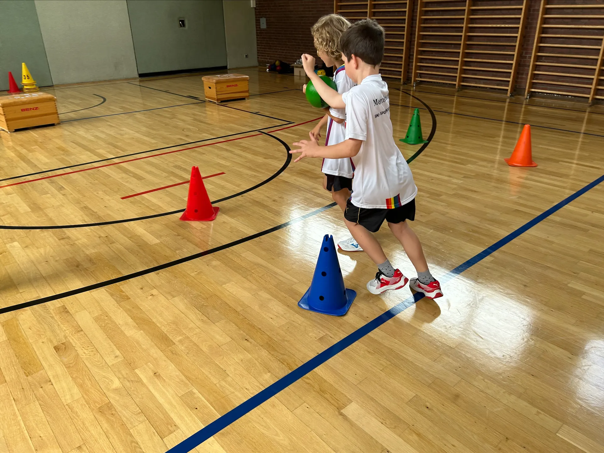 Zwei Kinder absolvieren mit einem Ball einen Parcours in einer Sporthalle.