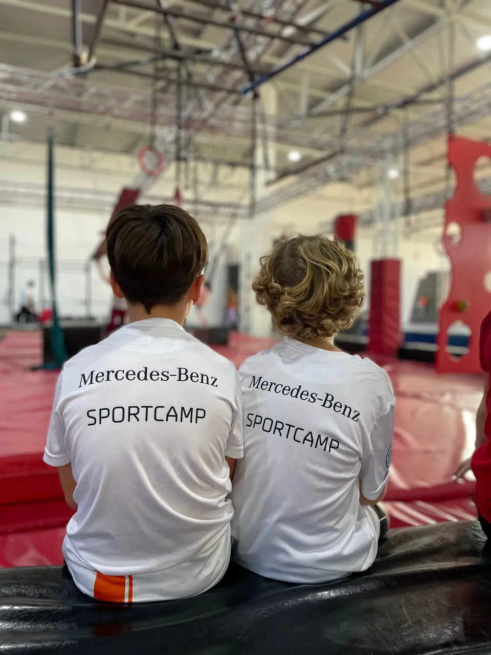 Zwei Kinder sitzen von hinten in einer Sporthalle und tragen Shirts mit der Aufschrift „Mercedes-Benz Sportcamp“.