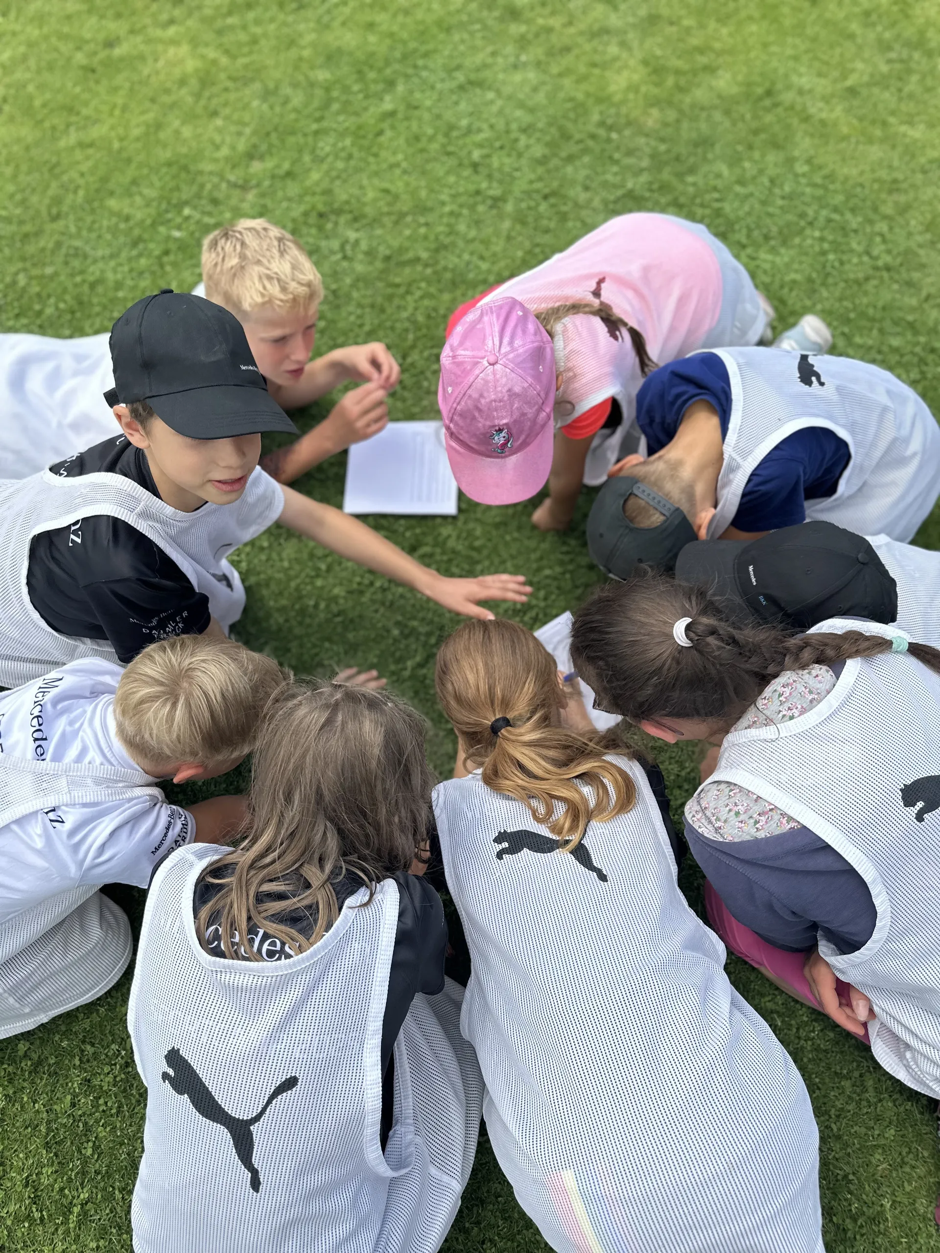 Kinder in Trainingswesten mit Puma-Logo sitzen im Kreis auf dem Rasen und schauen auf ein Papier.